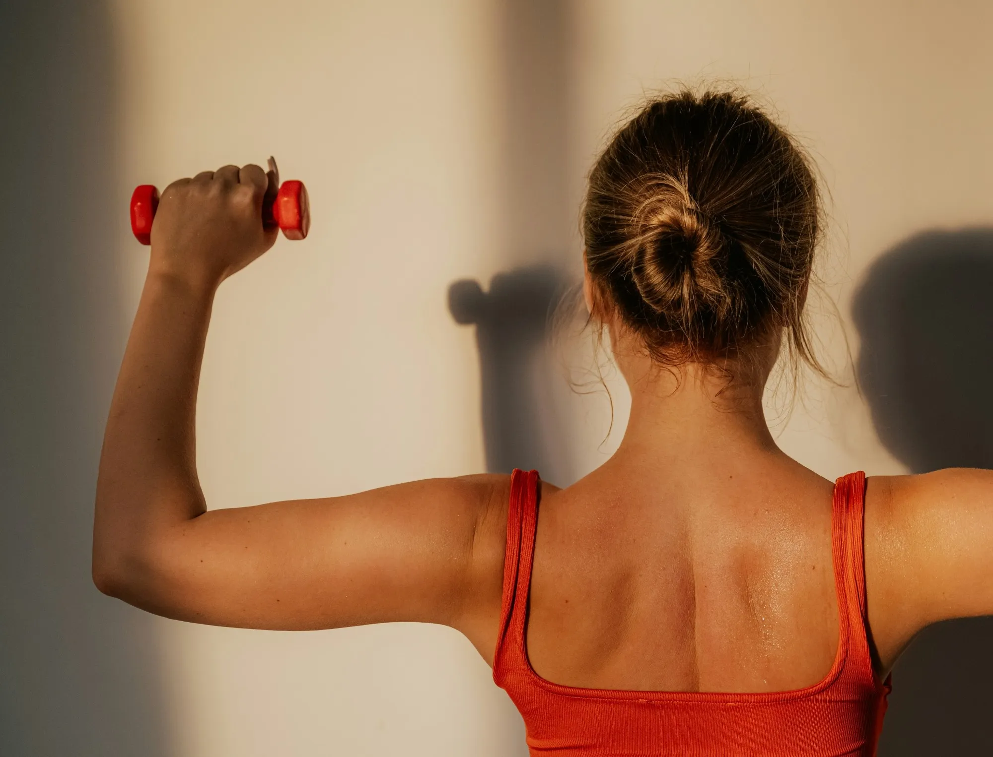 A woman in a red tank top lifts a small red dumbbell overhead in warm afternoon light